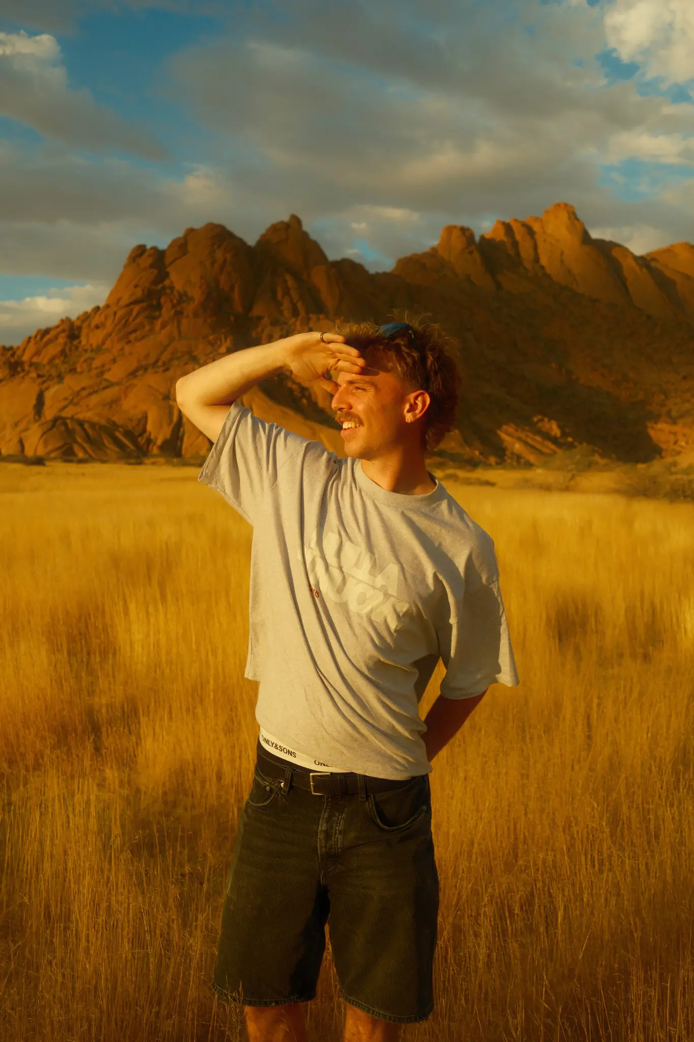 man shielding eyes in golden field with mountains at sunset